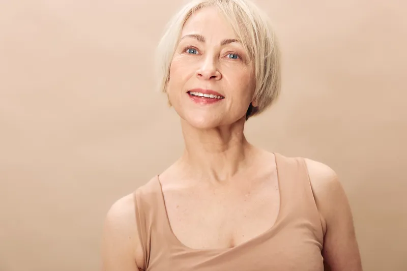 Portrait of a smiling mature woman with short blonde hair wearing a beige tank top against a neutral background. Concept of natural beauty, aging, confidence, and positive lifestyle.