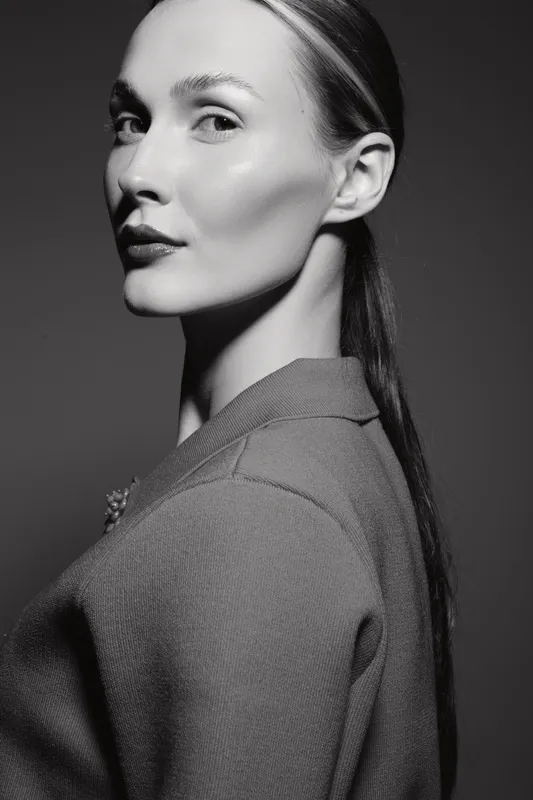 Profile of a young white European woman with fair skin and sleek hair in a textured jacket, studio portrait in black and white.