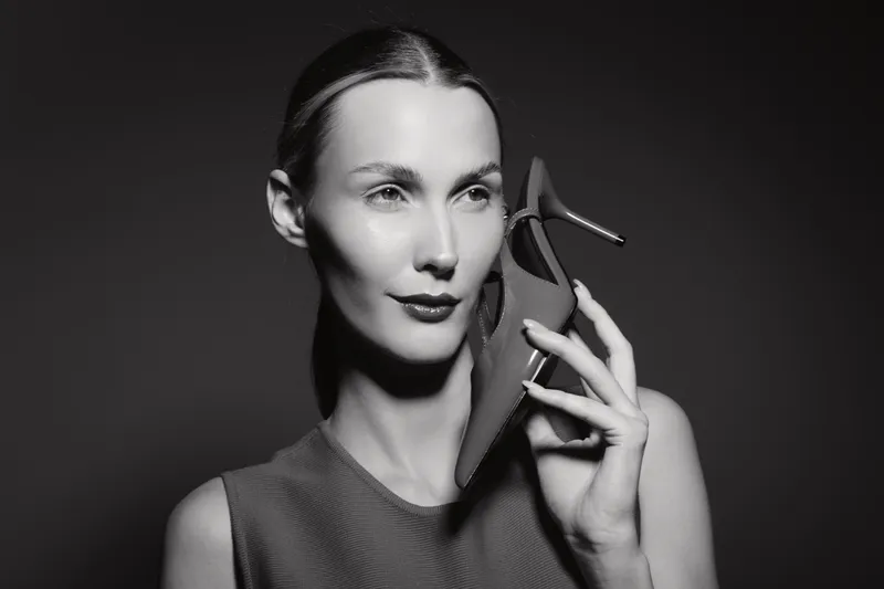 Elegant Caucasian woman with light skin holds high heel shoe near face against dark background. Concept for fashion and style.