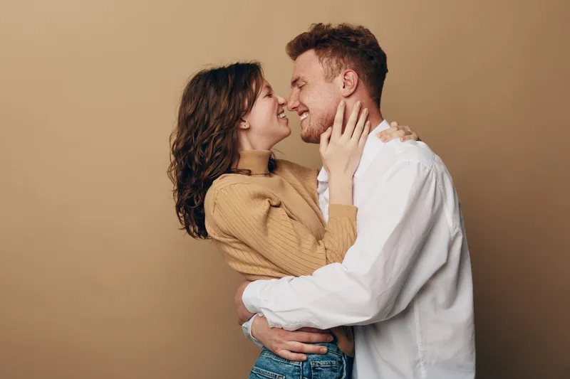 Happy young couple embraces and smiles looking at each other on beige background. Concept of love, relationship, affection, and romantic connection between man and woman.