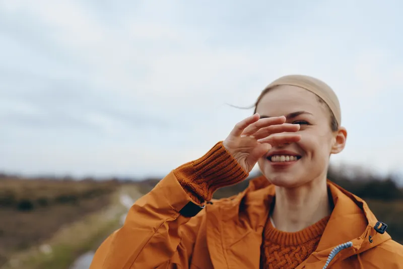 Smiling woman in an orange jacket and scarf outdoors in an autumn field, close portrait view, she covers her mouth with her hand, natural daylight, woman lifestyle, relaxed happy mood