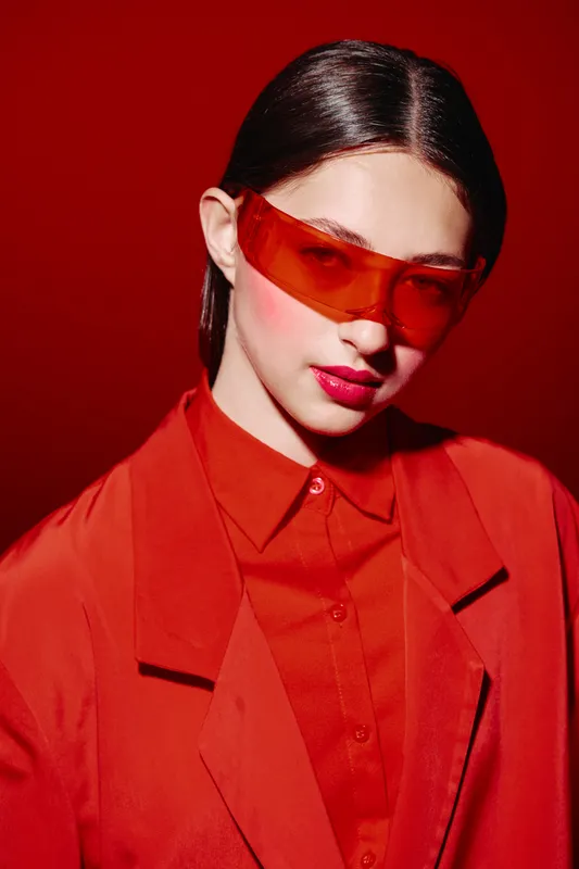 Young woman in vibrant red outfit and transparent orange sunglasses poses against red background. Modern beauty fashion concept featuring bold colors and stylish accessories.