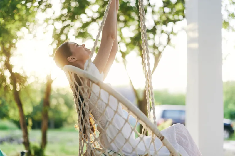 Serene woman enjoying a peaceful moment in a swing chair in a sunlit garden, wearing a white dress, evoking tranquility and joy