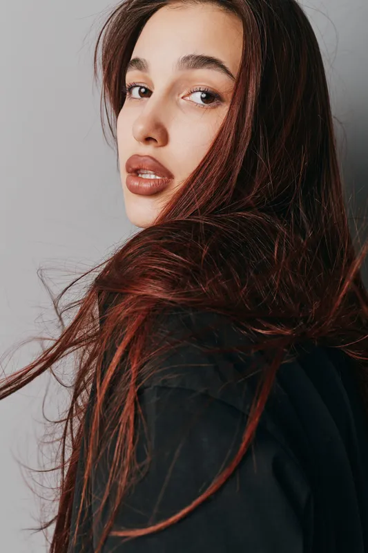 Portrait of young woman with long hair looking over shoulder against plain background. Closeup of female face with natural makeup and expressive eyes in casual clothing.