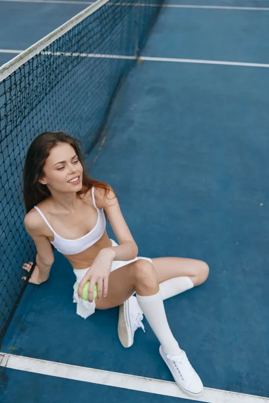 Young woman in athletic wear sitting on tennis court, smiling, holding tennis ball, showcasing healthy lifestyle, sports concept, vibrant blue background