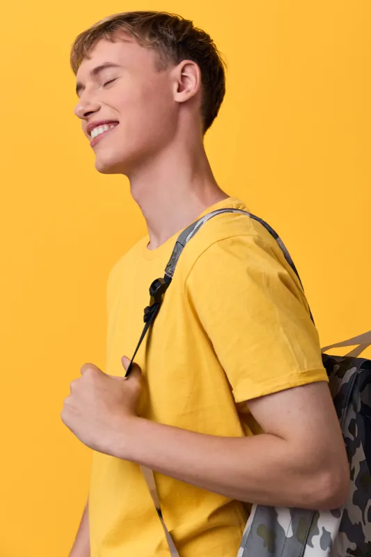 Youth wearing a yellow shirt with a backpack, smiling against a bright backdrop, showcasing casual fashion, energetic lifestyle, and carefree everyday mood.