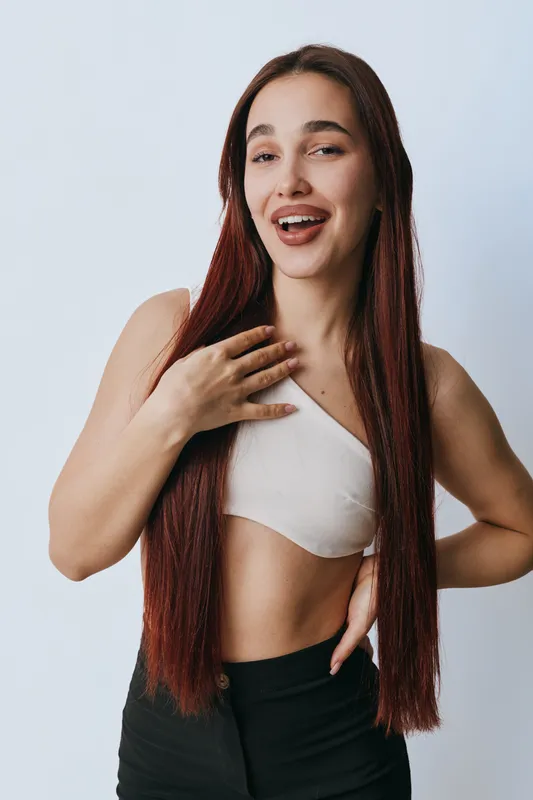 Young woman with long straight hair smiling and posing in casual outfit against white background. Female model with natural expression and confident posture for beauty concept.