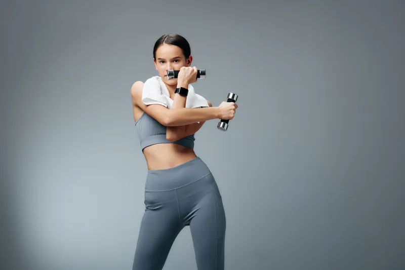Fitness sport woman in sleek gray activewear performs a dynamic workout with dumbbells, showing strength, balance and focus in a clean studio setting.