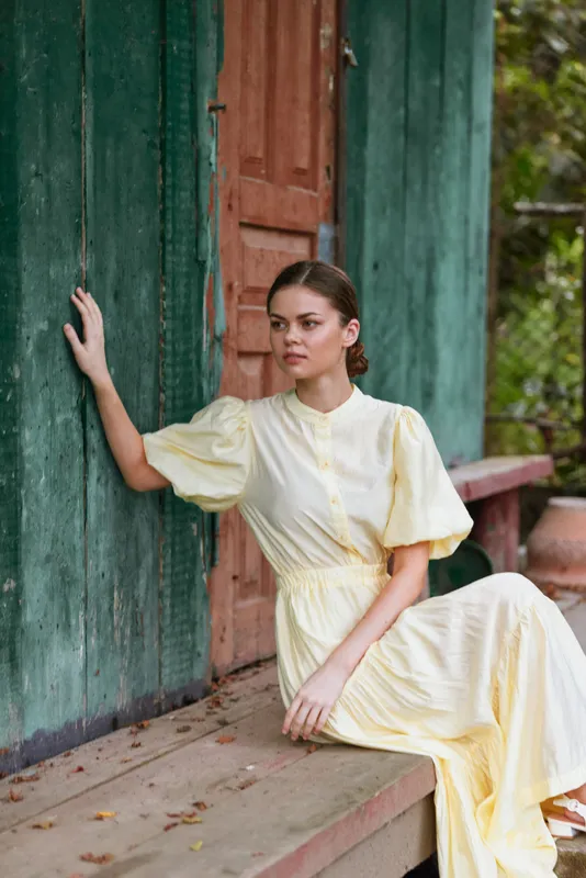 A woman in a pale yellow dress sits gracefully on a wooden porch beside a weathered doorway, exuding calm elegance and thoughtful poise under natural outdoor light
