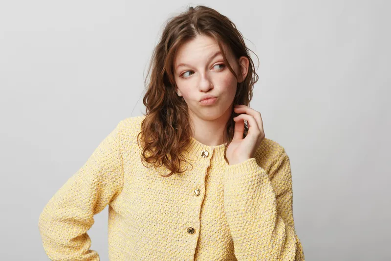 Young woman in yellow knitted sweater looks sideways with thoughtful expression on grey background. Concept for doubt, decision making, and contemplation in casual style portrait.