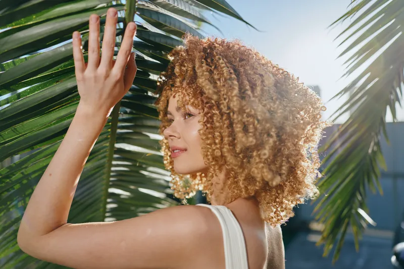 Curly woman with blonde hair touches palm leaves outdoors, dressed in white, enjoying sunlight and summer warmth, natural beauty in tropical green environment.