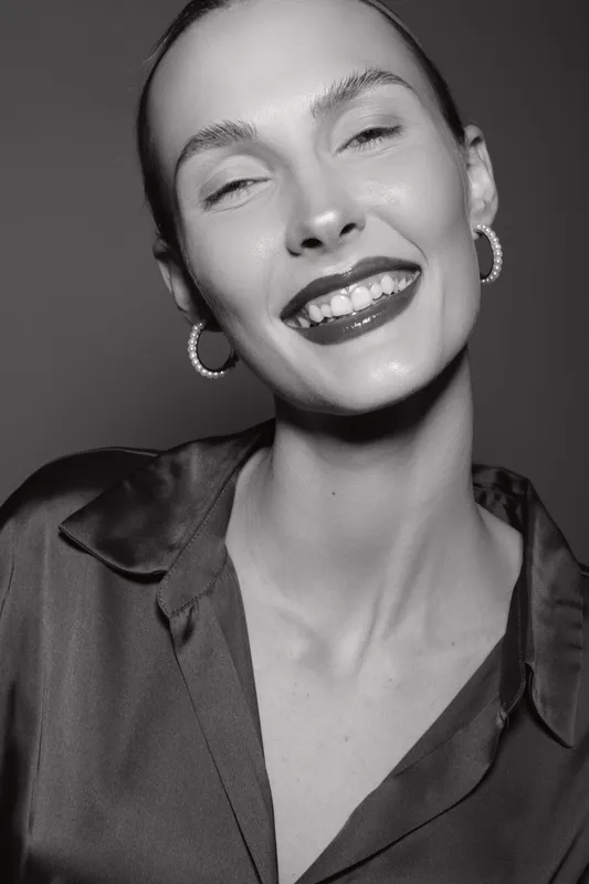 Smiling white woman with pale skin and slicked back hair wears shiny blouse and hoop earrings. Studio portrait with joyful expression, fashion, and beauty concept in black and white.