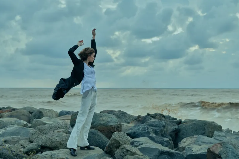 People in business attire on a rocky beach celebrate achievement, jacket flung, arms raised, wind in hair, sea horizon and dramatic clouds emphasizing success.