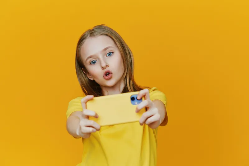 Cheerful girl taking a selfie with a yellow smartphone against a vibrant yellow background, capturing playful moments with bright colors