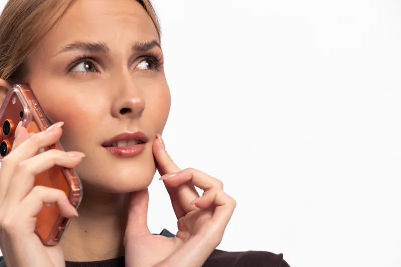 Young woman with natural makeup holding smartphone to ear looks worried and thoughtful isolated on white background. Concept for communication, concern, and decision making.