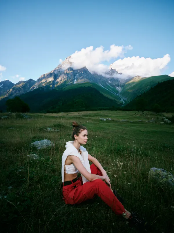 Woman sits in a green meadow at the foot of towering mountains, wearing red pants and a white top, gazing into the distance under a blue sky with fluffy clouds during outdoor travel