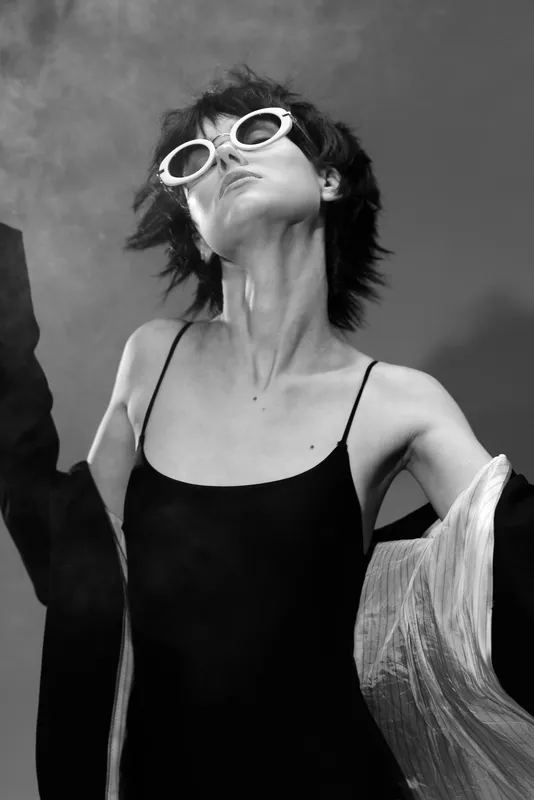 Black and white portrait of a stylish woman wearing sunglasses and a black dress with flowing fabric.