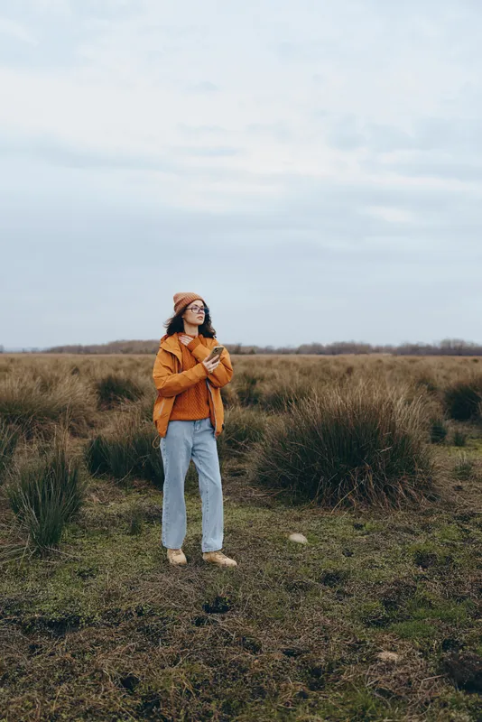 Woman lifestyle outdoors in a grassy field, standing in orange jacket and jeans, scarf and glasses, holding a smartphone, calm mood, cloudy sky, natural light.
