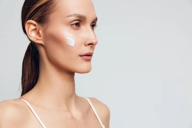 Young woman in close up profile applies face cream for skincare, smooth skin and beauty care, studio portrait with white background, calm expression, minimal makeup, hydration and glow.