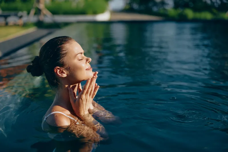 Young woman with wet hair enjoys a peaceful moment in an outdoor swimming pool under natural sunlight with closed eyes and relaxed expression.