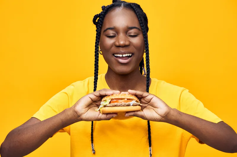 Young woman with braids joyfully holding a burger against a vibrant yellow background, representing healthy eating and happiness