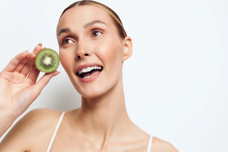 Happy woman holding fresh kiwi slice near face, smiling with open mouth, bright eyes, clean skin, white background, healthy fruit, natural vitamins, fresh lifestyle