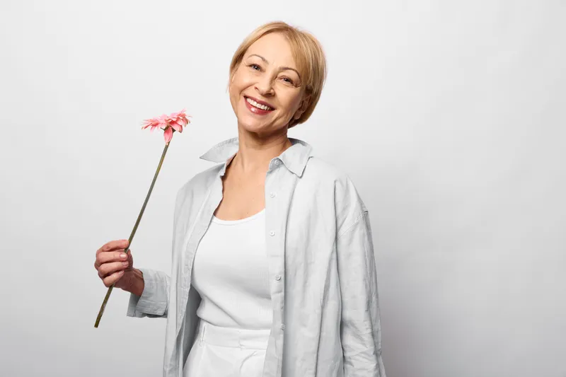 Smiling woman holds a pink flower in a studio portrait on a clean white background, spring mood for female beauty, soft light adds joy