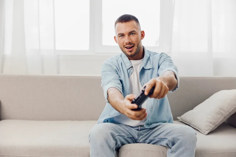 Engaged man playing video games on a couch, showcasing excitement and leisure. The bright indoor setting enhances the vibrant atmosphere, making it ideal for lifestyle content.