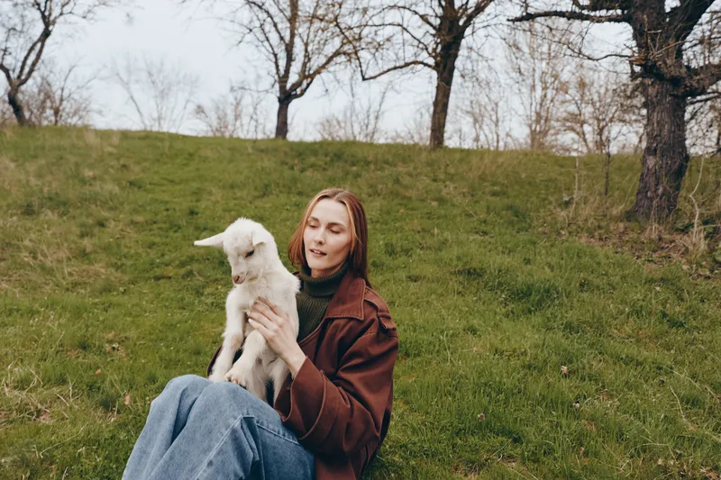 Woman sits on a grassy hill holding a white goat kid, enjoying outdoor lifestyle, gentle serene moment of companionship in a parklike countryside setting, calm and warm