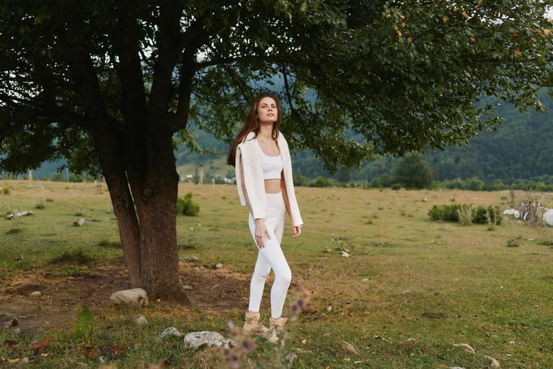 Young woman in white outfit standing under large tree in open field with mountains in background natural light peaceful atmosphere casual lifestyle