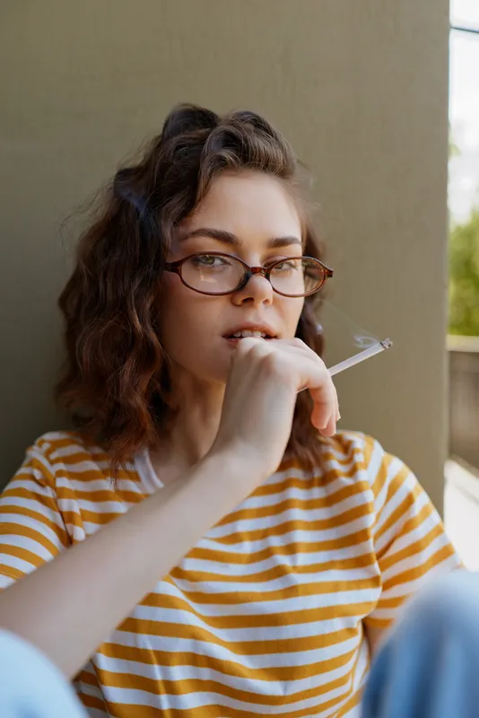 Smiling woman with striped shirt and glasses relaxing on a balcony, holding a cigarette near her lips. Casual lifestyle portrait emphasizing mood, confidence, and everyday relaxation.