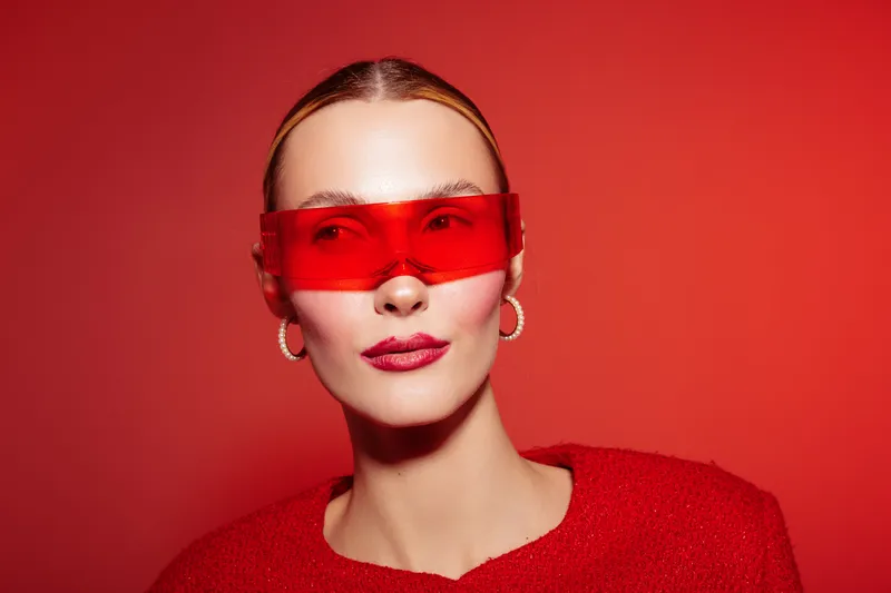 Stylish woman with red sunglasses and red sweater poses against red background. Fashion portrait with modern accessories and confident expression. Concept of bold style and color.
