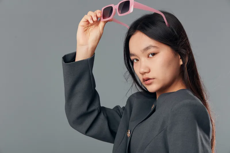 Young woman with long dark hair is holding pink sunglasses above her head wearing a dark gray blazer posing on neutral gray background in studio portrait fashion style.