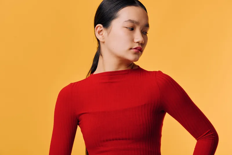 Young woman with dark hair in ponytail wearing red long sleeve top posing looking sideways on yellow background. Casual fashion portrait with serious expression and natural makeup.