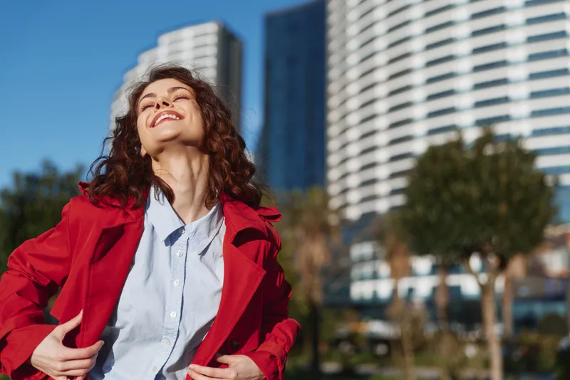 Young woman with curly brown hair smiles brightly in a red jacket and light blue shirt in a sunlit city park, with an urban backdrop suggesting freedom and confidence