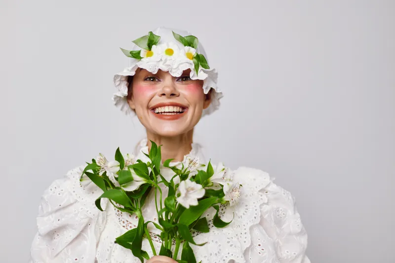 Cheerful woman in floral headwear, holding fresh flowers, wearing a vintage white dress, capturing joy and beauty in a bright and simple background