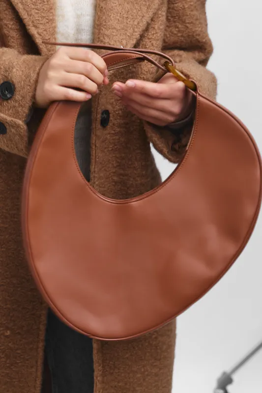 asian woman wearing a brown coat holds a circular brown handbag in a chic street setting, presenting a stylish and modern accessory with subtle elegance and poised posture.