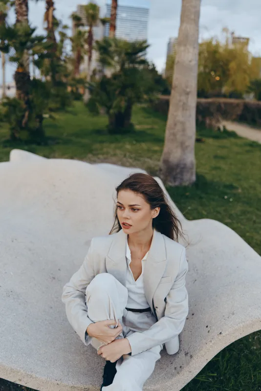 Woman in light suit sitting outside on abstract stone bench with contemplative expression in spring park setting