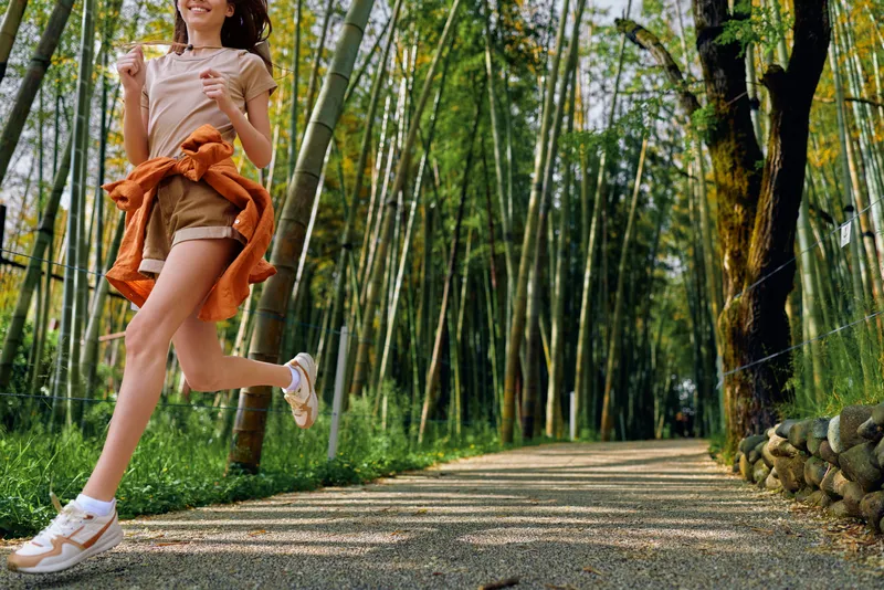 Woman running in bamboo forest on a sunlit trail, wearing sneakers and summer shorts, motion capture of active outdoor fitness and joyful jogging among tall green stalks.