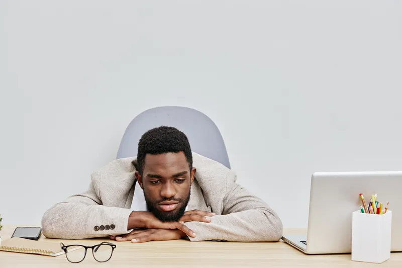 Focused young african-american man with beard resting head on folded arms at desk in office workspace, looking bored or tired, surrounded by laptop, glasses, and office supplies, professional workplac