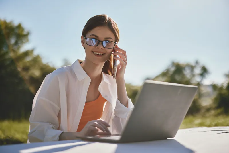 Young woman working on a laptop outdoors, smiling and wearing sunglasses, reflecting a positive and productive lifestyle in nature