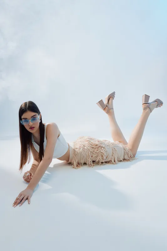 Young woman in stylish outfit and sunglasses lies on white surface under bright light. Concept of woman beauty fashion with modern accessories and summer style.