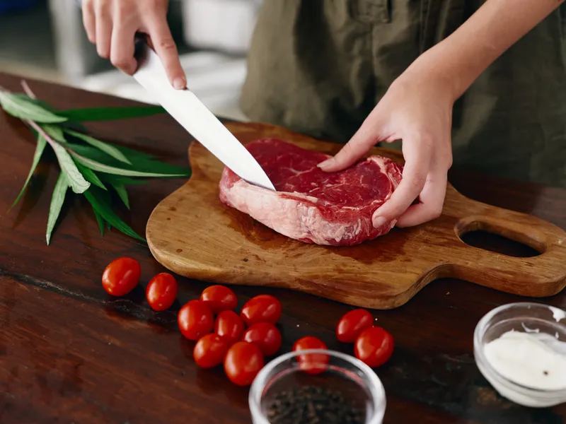 Женщина с ножом в руке нарезает свежее стейковое мясо для жарки на кухне
