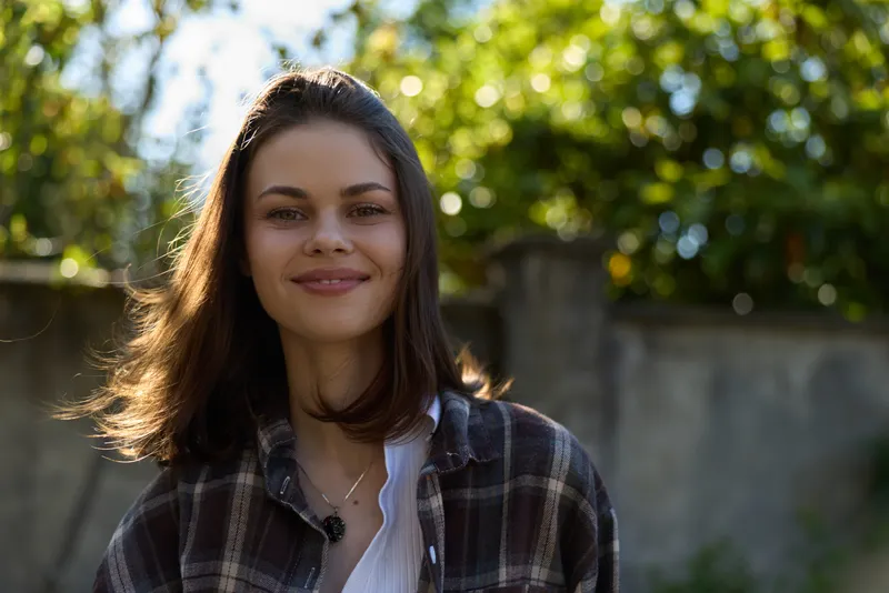 Smiling woman outdoors in a plaid shirt, head and shoulders portrait against a sunlit park background, showcasing casual lifestyle, confidence, and natural warmth