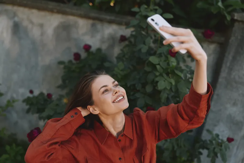Smiling woman in a red shirt taking a selfie outdoors with a smartphone. Bright garden setting conveys casual lifestyle, happiness, and personal creativity. Natural light highlights a cheerful moment