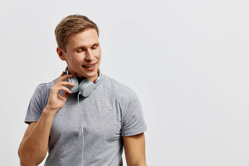 Smiling young Caucasian man wearing casual gray t-shirt, listening to headphones, holding a phone, seen against a plain white background, studio portrait, lifestyle, cheerful, relaxed, modern style.