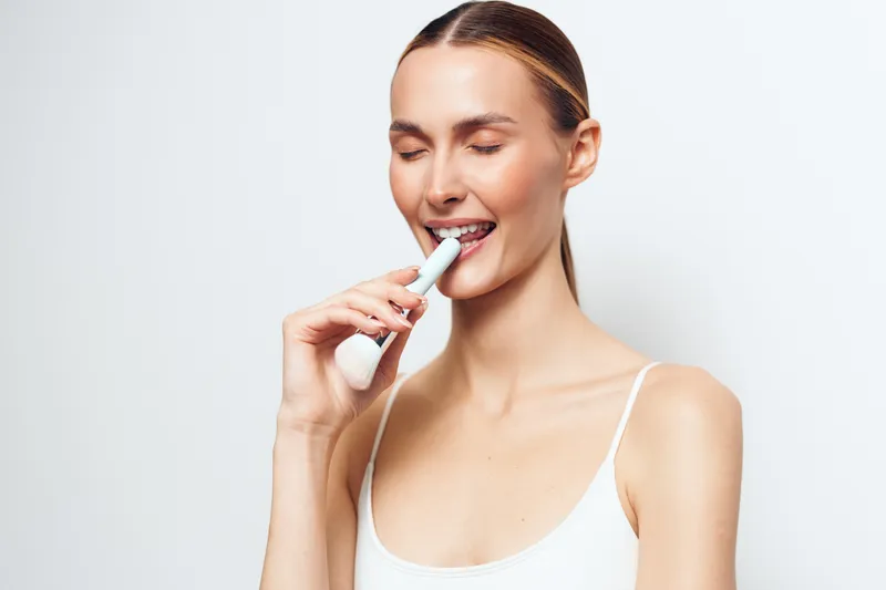 Young woman with closed eyes brushing teeth with electric toothbrush on white background. Concept of dental care, hygiene, and morning routine with soft natural lighting.
