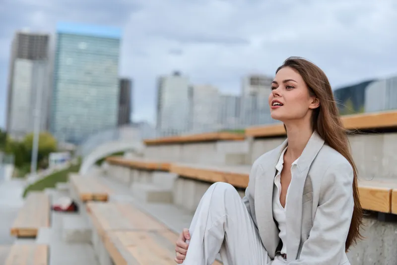 Young woman in light business suit with confident expression sitting on outdoor wooden steps in modern urban environment cityscape