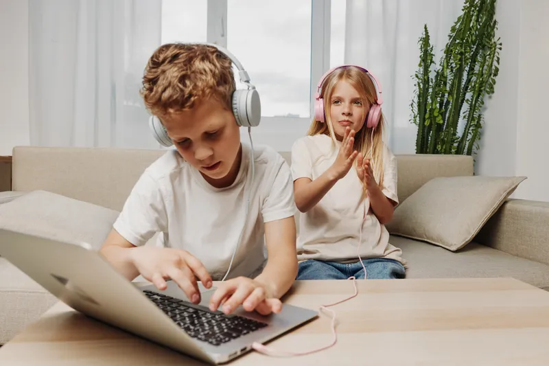 Focused boy with headphones uses laptop while girl claps hands sitting on couch in modern living room. Concept of technology, childhood, learning, and music entertainment.