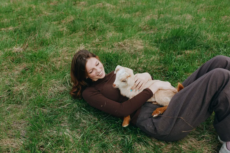 A woman lies on grass outdoors, smiling while embracing a small dog. The scene conveys warmth, companionship, and relaxation in a casual lifestyle moment.